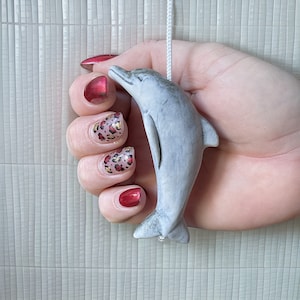 Op de afbeelding: Een hand met rode en gouden dierenprint nagellak houdt een grijze keramische dolfijn trekkoord vast.