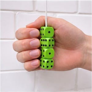 Pode incluir: Uma pilha de quatro dados verde brilhante, cada um com pontos pretos, suspensos por uma corda branca. Os dados são segurados em uma mão contra um fundo de azulejos brancos. Um divertido acessório de jogo, cada dado mede cerca de 2,5 cm.