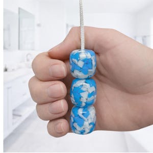 May include: A light pull with three cube-shaped beads in shades of blue and white. The beads are strung on a white cord. The background is a blurred white bathroom, suggesting a bathroom accessory.