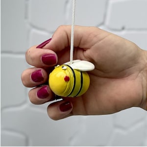 Può includere: Un ornamento a forma di ape gialla con strisce nere, ali bianche e guance rosse, sospeso da un filo bianco. L'ape è tenuta in una mano con smalto rosso scuro. Lo sfondo è un muro bianco strutturato.