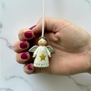 Könnte beinhalten: Kleine Keramik-Engelsfigur mit einem Stern auf der Brust. Der Engel hat ein weißes Gewand, grüne Flügel und eine Schnur zum Aufhängen. Die Figur wird von einer Hand mit rotem Nagellack gehalten.