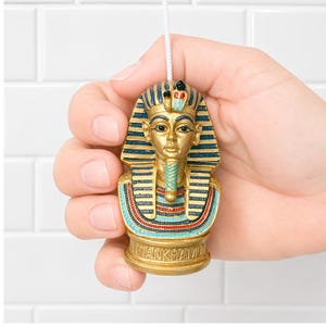 May include: A gold-colored bust of Tutankhamun, with blue and red accents, held against a white tile background. The bust features a detailed headdress and collar. The base of the bust reads "Tutankhamun". A white string is attached to the top.