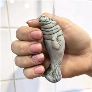 May include: A gray manatee figurine with dark gray stripes and spots, suspended by a silver cord. The manatee is held in a hand with pink nail polish.