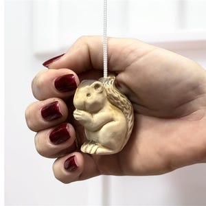 May include: A light brown squirrel-shaped pull chain for a light fixture. The squirrel is holding a nut and has a textured tail. The pull chain is attached to a white cord.