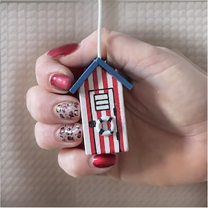 May include: A small wooden beach hut decoration, featuring red and white stripes, a blue roof, and a white life preserver. It is suspended by a white string.