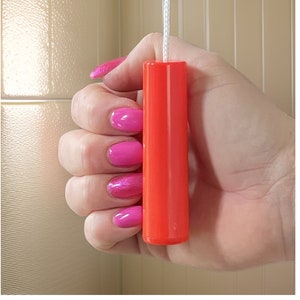 Puede incluir: Un cordón de tiro de plástico naranja brillante para una lámpara. El cordón está siendo sujetado por una mano con esmalte de uñas rosa.