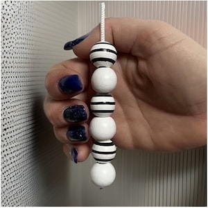 Puede incluir: Una cadena de cuentas blancas con rayas negras, colgando de un cordón blanco. La cadena de cuentas está sujeta en una mano con esmalte de uñas azul oscuro. Las cuentas alternan entre blanco sólido y blanco con rayas negras.