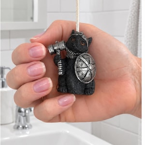 May include: A black cat figurine light pull featuring a warrior theme with a silver-coloured helmet, a silver-coloured shield, and a silver-coloured hammer held in one paw. The cat has orange eyes and is attached to a white cord for ceiling light operation.