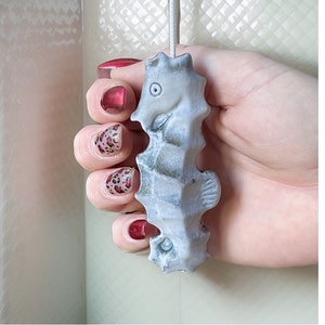 May include: A light gray seahorse-shaped candle with a textured surface. The candle is held by a hand with red and leopard print nail polish. The seahorse has detailed fins and a small eye.