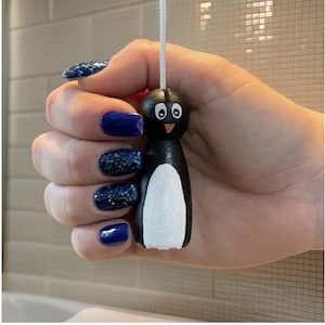 Può includere: Tirante per la luce a forma di pinguino bianco e nero con becco arancione, appeso a un cordino bianco. La mano che tira il tirante ha lo smalto blu.