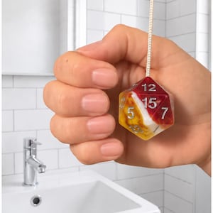 May include: A light pull cord featuring a multi-coloured icosahedron-shaped die as a handle. The die has a marbled pattern in shades of red, white, and yellow with silver-coloured numbers, including 12, 15, 5, and 7 clearly visible on the faces.