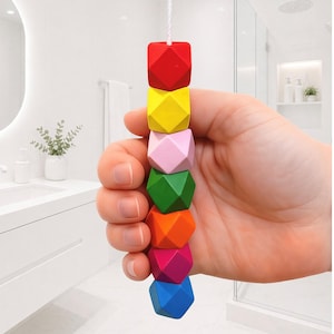 May include: A hand holds a string of colorful, geometric wooden beads. The beads are in a rainbow of colors: red, yellow, pink, green, orange, magenta, and blue. The background is a white bathroom.
