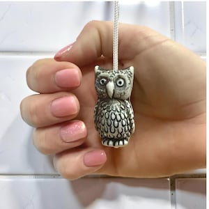 以下が含まれることがあります： 紐の付いた、小さく詳細なフクロウの置物。フクロウは主にグレーで、白いアクセントとテクスチャ加工された表面があります。 大きな丸い目とくちばしがあります。 置物は白いタイルの背景に対して保持されています。