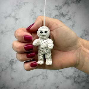Può includere: Piccola statuetta di mummia bianca con occhi neri e bende avvolte, sospesa da un filo bianco. Lo sfondo è un grigio e bianco screziato.