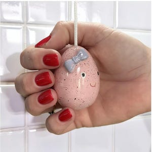 Könnte beinhalten: Ein rosa, gesprenkeltes, eiförmiges Ornament mit hellblauer Schleife und lächelndem Gesicht. Das Ornament wird an einer weißen Schnur gehalten. Die Hand, die es hält, hat roten Nagellack. Hintergrund: weiße Fliesenwand.