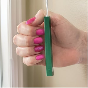 May include: A green plastic window blind pull cord with a white string attached. The cord is being held by a hand with pink nail polish.