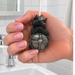 May include: A light pull featuring a small, dark grey, sculpted creature wearing a helmet and holding a round, silver-coloured shield with a cross design. The figure is attached to a white cord, showcasing detailed textures on the armour and shield.