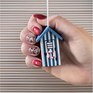 Op de afbeelding: Een miniatuur houten strandhuisornament met blauwe en witte strepen, een blauw dak en een rood-witte reddingsboei. Het ornament wordt door een hand tegen een gestreepte achtergrond gehouden.