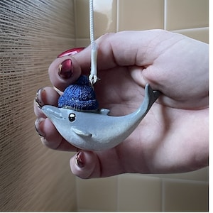 May include: A gray dolphin figurine wearing a blue knit hat. The dolphin is hanging from a white string.