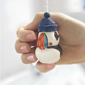 Op de afbeelding: Een kleine papegaaiduikerfiguur met een blauwe gebreide muts. De papegaaiduiker heeft een wit lichaam, zwarte accenten en een kleurrijke snavel. De figuur wordt in een hand gehouden tegen een vage achtergrond.