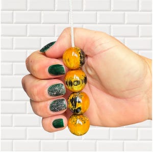 Op de afbeelding: Een decoratieve trekker met vier oranje en zwarte harskralen aan een wit koord. De kralen worden vastgehouden in een hand met groene en glitterende nagellak. De achtergrond is een witte tegelmuur.