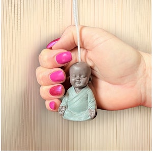 May include: A small, meditating figurine of a Buddha in a seated position. The figurine is made of a light gray material and has a serene expression on its face. The figurine is being held in a hand with pink nail polish.