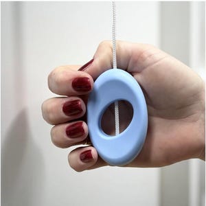 May include: A pale blue oval pull with a white cord, held in a hand with red nail polish. The pull has a central opening and is made of a smooth material. The cord is thin and white.