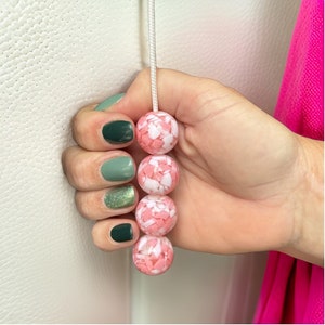 Puede incluir: Una cadena de cuentas rosadas y blancas con un patrón de mármol. Las cuentas están enhebradas en un cordón blanco y se sostienen en una mano con esmalte de uñas verde.