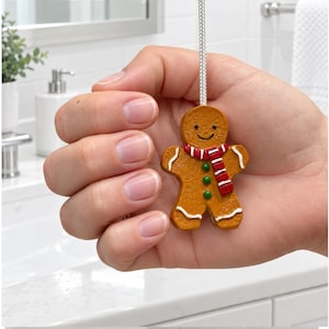 Könnte beinhalten: Ein Lichtzug in Form eines Lebkuchenmanns, bestehend aus einer braunen Keksfigur mit lächelndem Gesicht, einem rot-weiß gestreiften Schal und drei grünen Knöpfen. Die Figur ist mit weißem Zuckerguss verziert und hängt an einer silbernen Schnur.