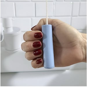 以下が含まれることがあります： 白い紐が付いた水色のトイレのフラッシュハンドルが、赤いマニキュアを塗った手で持たれています。ハンドルは円筒形で、下に向かってわずかに細くなっています。背景は白いタイルの壁と白いトイレです。