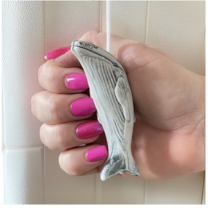 Op de afbeelding: Een walvisvormige, wit en grijze keramische trekker met een wit koord. De trekker wordt vastgehouden in een hand met felroze nagellak. De walvis heeft gedetailleerde markeringen en een textuuroppervlak.