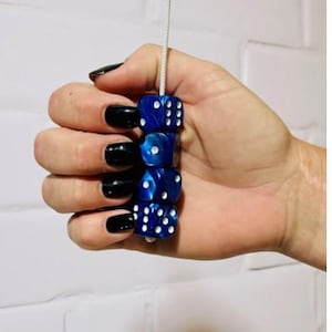 May include: Four blue dice with white dots, strung on a white cord. The dice are held in a hand with black nail polish. The dice are a deep blue colour. The image is set against a white background.