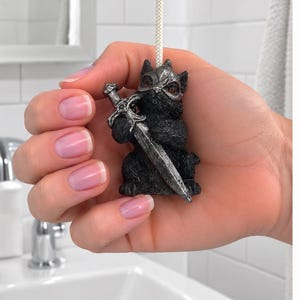 May include: A black cat light pull featuring a silver-colored helmet and a silver-colored sword held in its paws. The cat figure is attached to a white cord, designed for use as a decorative handle for a light switch or ceiling fan pull chain.