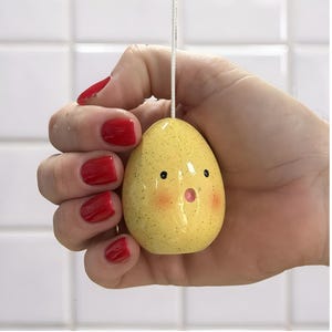 Könnte beinhalten: Ein gelbes, gesprenkeltes, eiförmiges Ornament mit überraschtem Gesichtsausdruck. Das Ornament hat schwarze Augen, rosige Wangen und einen offenen, rosa Mund. Oben ist eine weiße Schnur befestigt. Der Hintergrund ist eine weiße geflieste Wand.