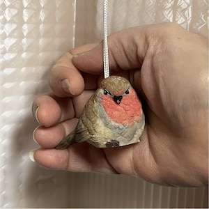 May include: A small, brown and gray bird figurine with a red breast. The bird is holding a white string in its beak and is being held in a hand.