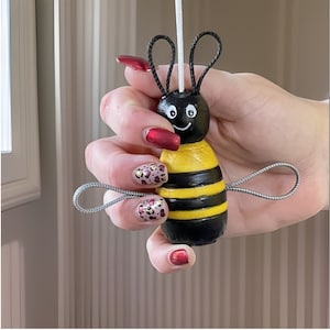 Puede incluir: Adorno de abeja pintado a mano. La abeja de madera tiene un cuerpo a rayas negro y amarillo, una cara sonriente y antenas y alas de alambre. Tiene un cordón blanco para colgar.