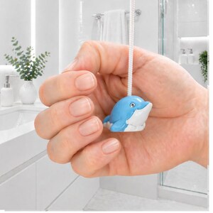 Puede incluir: Tirador de luz con forma de delfín azul y blanco con un cordón blanco. El delfín tiene una cara sonriente y se sostiene frente a un baño blanco. El baño tiene un lavabo blanco, un jarrón blanco con plantas verdes y una ducha.