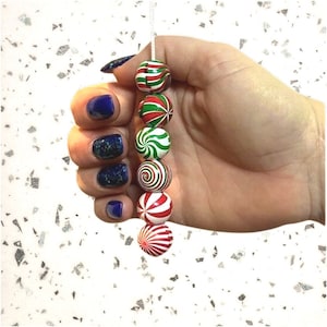 Peut inclure: Une chaîne de six boules de Noël rondes et colorées tenues dans une main. Les boules présentent des motifs rouges, verts et blancs, notamment des rayures et des spirales. La main a du vernis à ongles bleu.