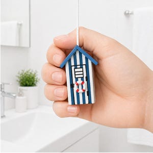 Op de afbeelding: Een kleine blauw-wit gestreepte strandhuisje lichtschakelaar met een rood-witte reddingsboei. De lichtschakelaar wordt door een hand vastgehouden voor een witte badkameromgeving.