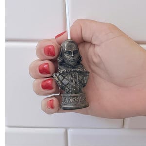May include: A small, antique-style bust of William Shakespeare, held against a white background. The bust is a dark metallic color and features intricate details of Shakespeare's face and clothing. The base of the bust reads "Shakespeare".