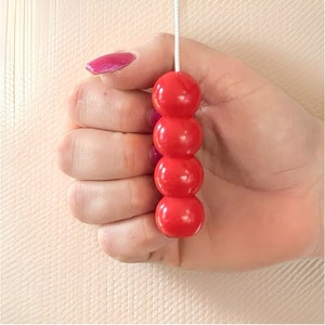 Könnte beinhalten: Eine rote Perlen-Zugkette für eine Leuchte oder einen Rollladen. Die Kette besteht aus vier runden, glänzenden roten Perlen, die auf einer weißen Schnur aufgereiht sind. Die Perlen werden in einer Hand mit pinkfarbenem Nagellack gehalten.
