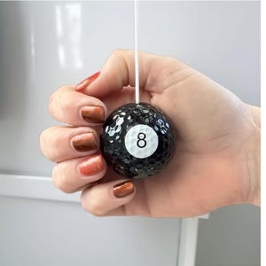 Könnte beinhalten: Eine schwarze Billardkugel mit der Nummer 8 auf einem weißen Kreis, aufgehängt an einer weißen Schnur. Der Ball hat eine strukturierte Oberfläche. Das Bild ist ein dekoratives Objekt.