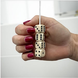 May include: Four ivory dice with black pips are strung together on a white cord. The dice are held in a hand with red nail polish. The dice are a classic game accessory.