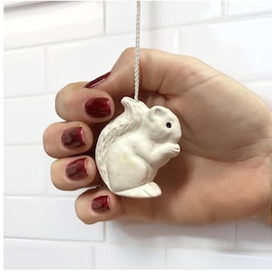 May include: A white ceramic squirrel light pull with a small black eye, suspended by a white cord. The squirrel is in a sitting position, holding something in its paws. The background is a white tiled wall.