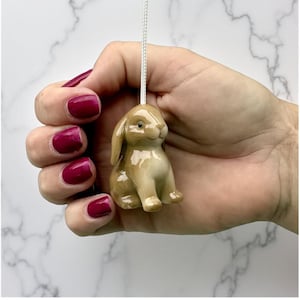 Puede incluir: Una pequeña figura de conejo de cerámica de color marrón claro, suspendida por un cordón blanco. El conejo se sostiene en una mano con esmalte de uñas rojo oscuro. La figura tiene un acabado brillante y presenta pequeñas patas y una cara sonriente.