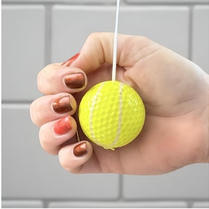 May include: A bright yellow golf ball-shaped light pull with a white cord. The golf ball has a dimpled surface and is held in a hand with polished nails. The background is a white tiled wall.