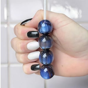 May include: Four blue and black glass beads on a white cord. The beads are held in a hand with black and white nail polish. The beads have a glossy finish and reflect light.