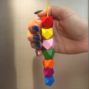 Könnte beinhalten: Eine Kette aus bunten Holzperlen in geometrischen Formen, darunter Rot, Gelb, Rosa, Grün, Orange, Lila und Blau. Die Perlen sind auf einer gelben Schnur aufgefädelt. Die Kette wird in einer Hand mit blauem Nagellack gehalten.