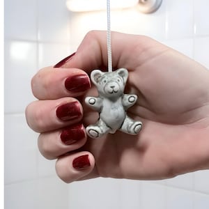 May include: A small, gray teddy bear light pull with a white cord. The bear has a smiling face and outstretched arms. The background is a white tiled wall. The hand holding the pull has dark red nail polish.
