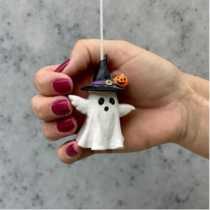 Può includere: Una statuetta di fantasma bianco con un cappello da strega nero con una fascia viola e una piccola zucca arancione. Il fantasma ha occhi neri e una bocca piccola. È sospeso da un filo bianco, ideale per Halloween.
